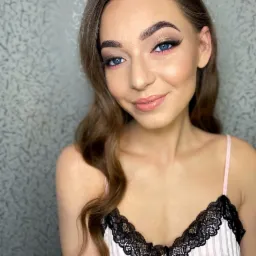 Portret uśmiechniętej kobiety z profesjonalnym wizażem: złote cienie, różowy akcent pod okiem, podkreślone brwi i błyszczące usta. Delikatne fale na włosach.