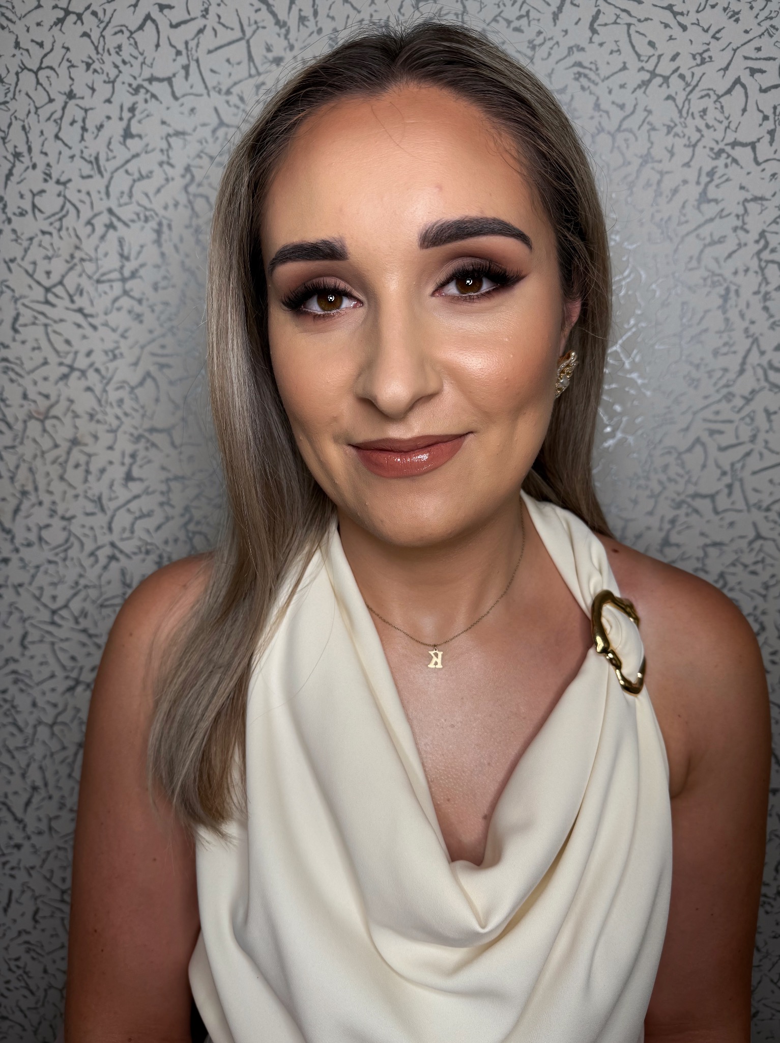 Portret kobiety z profesjonalnie wykonanym makijażem, uwydatniającym oczy i usta, z delikatną biżuterią i blond włosami na tle wzorzystej ściany.