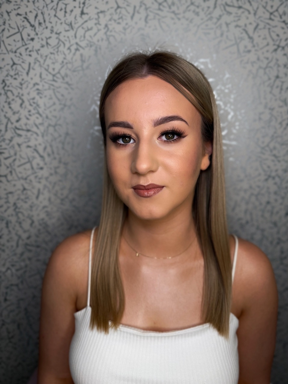 Portret kobiety z delikatnym makijażem, podkreślone oczy i usta w odcieniach brązu, gładkie blond włosy na tle szarej ściany.