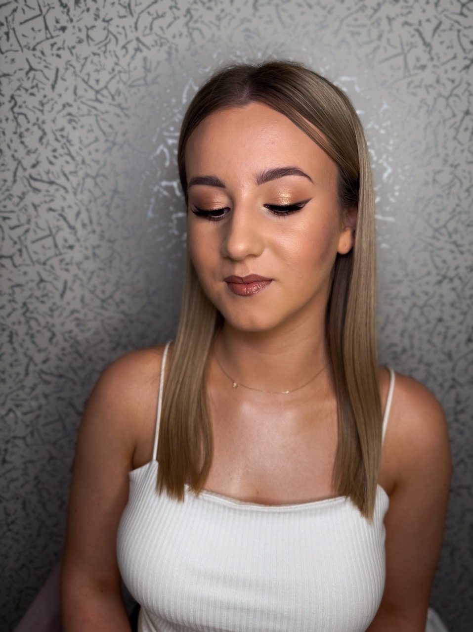 Portret kobiety z delikatnym, złotym makijażem oczu, czarną kreską i błyszczącymi ustami. Uwagę zwraca precyzyjny kontur i stonowana kolorystyka.