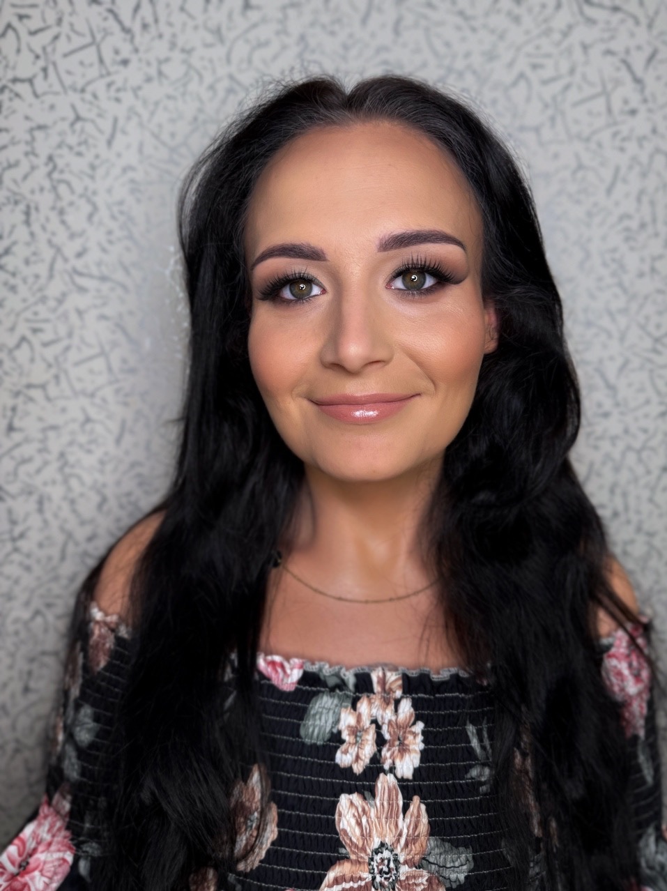 Portret kobiety z delikatnym, profesjonalnym make-up'em, podkreślającym oczy i usta, z długimi, ciemnymi włosami i w bluzce w kwiaty na tle wzorzystej ściany.