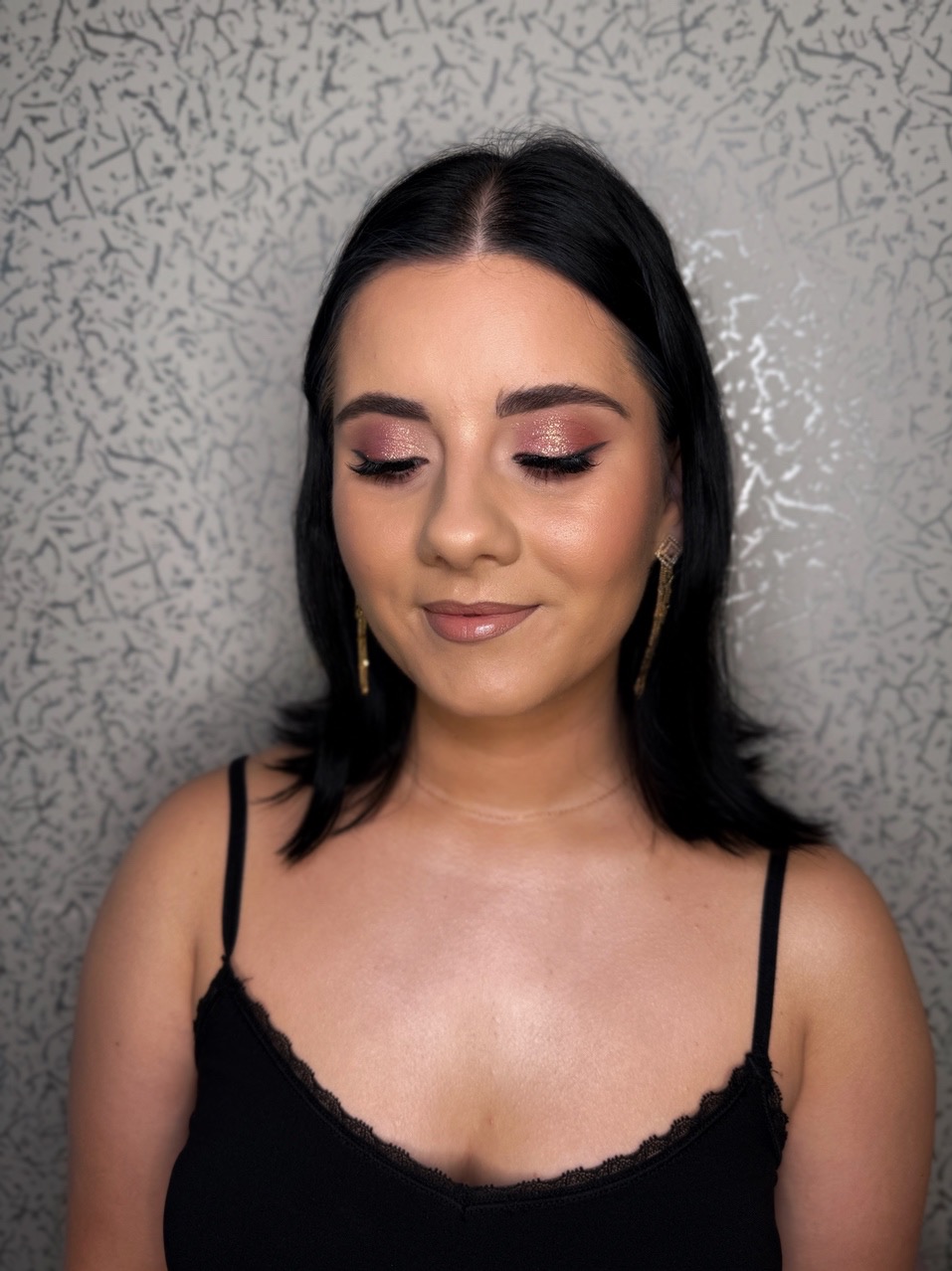 Portret kobiety z zamkniętymi oczami, prezentującej wieczorowy look: różowe cienie z brokatem i delikatny kontur. Długie kolczyki i czarna bluzka na ramiączkach.