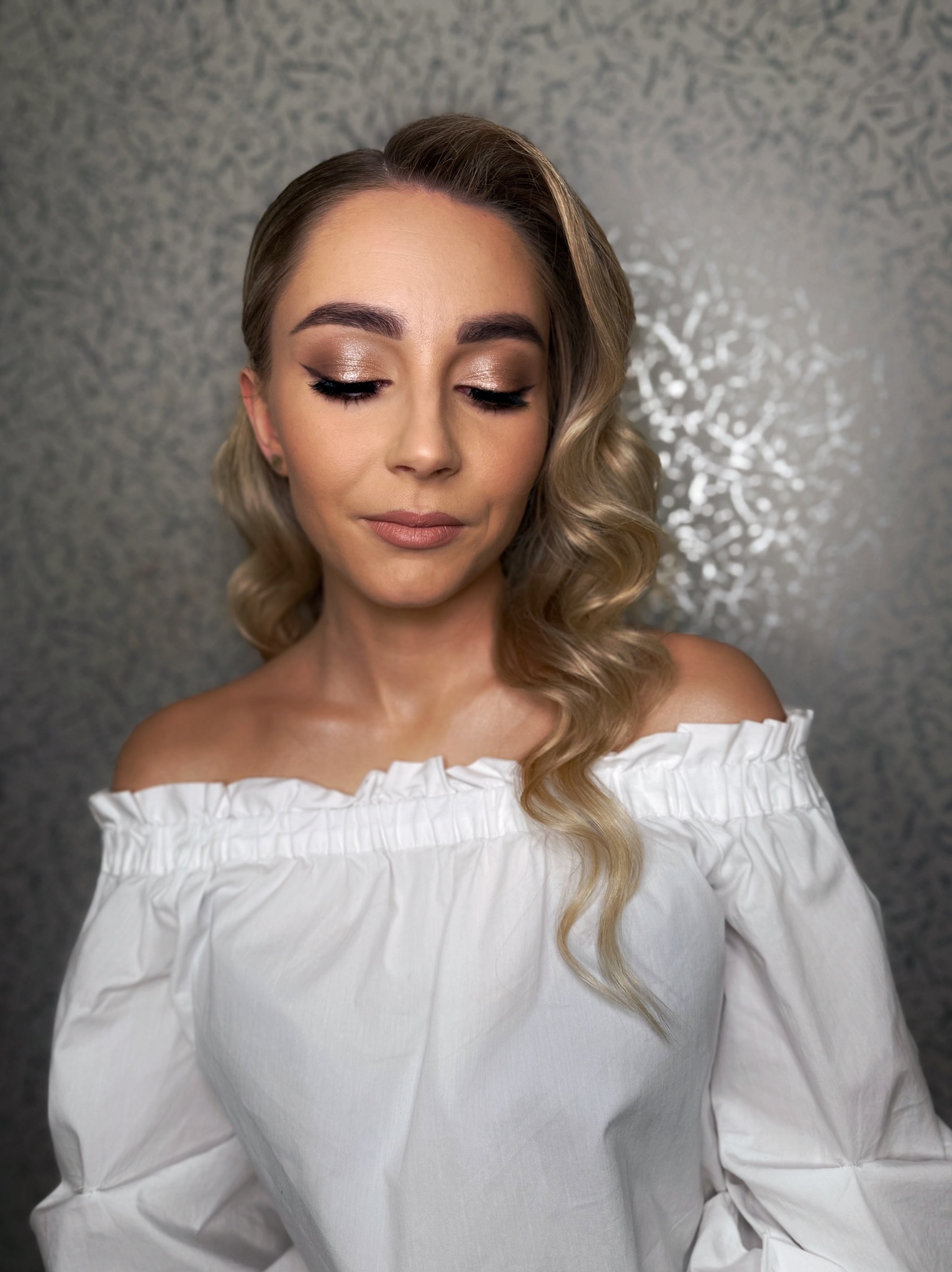 Portret blondynki z zamkniętymi oczami, prezentującej złoty makijaż oczu i delikatny makijaż ust, w białej bluzce z odkrytymi ramionami na tle srebrnej ściany.