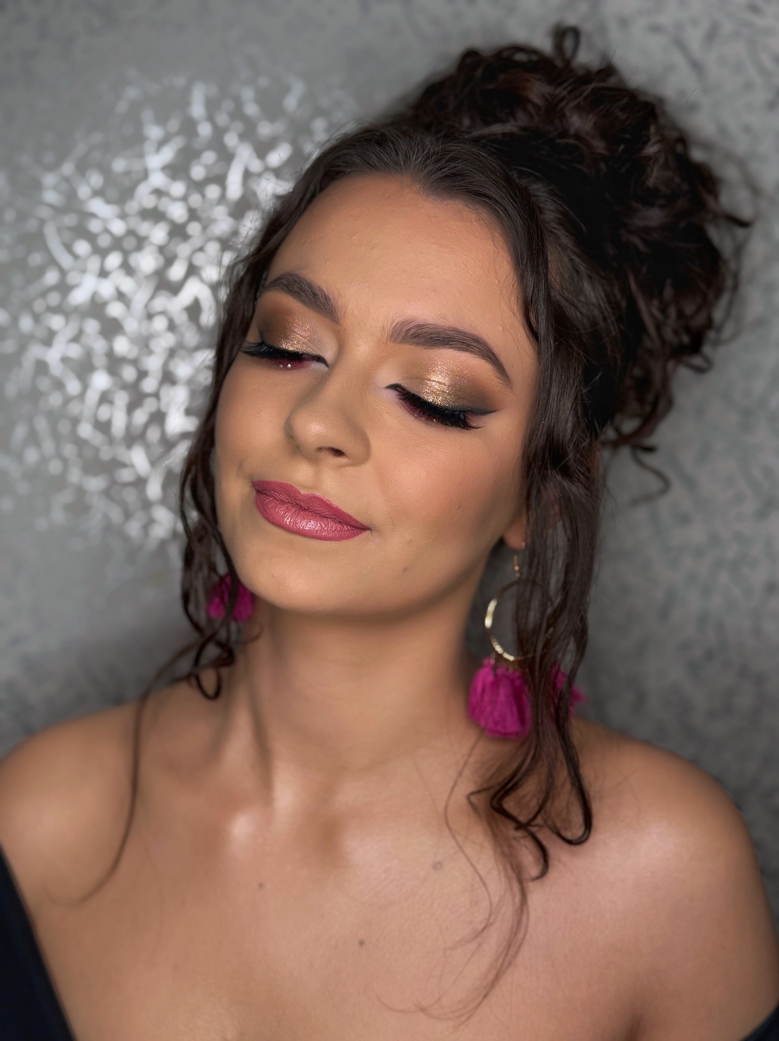 Kobieta z zamkniętymi oczami prezentuje złoty makijaż powiek i różowe usta, z upiętymi włosami i różowymi kolczykami na tle srebrzystej ściany.