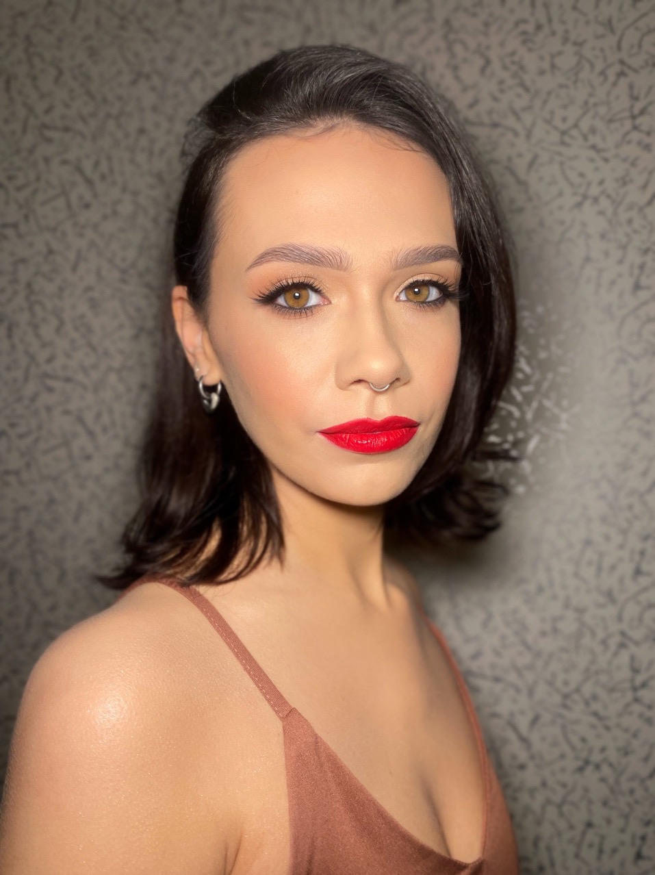 Portret kobiety z wyrazistym makijażem: czerwone usta, podkreślone oczy i brwi. Piercing w nosie, srebrne kolczyki. Krótkie, ciemne włosy.