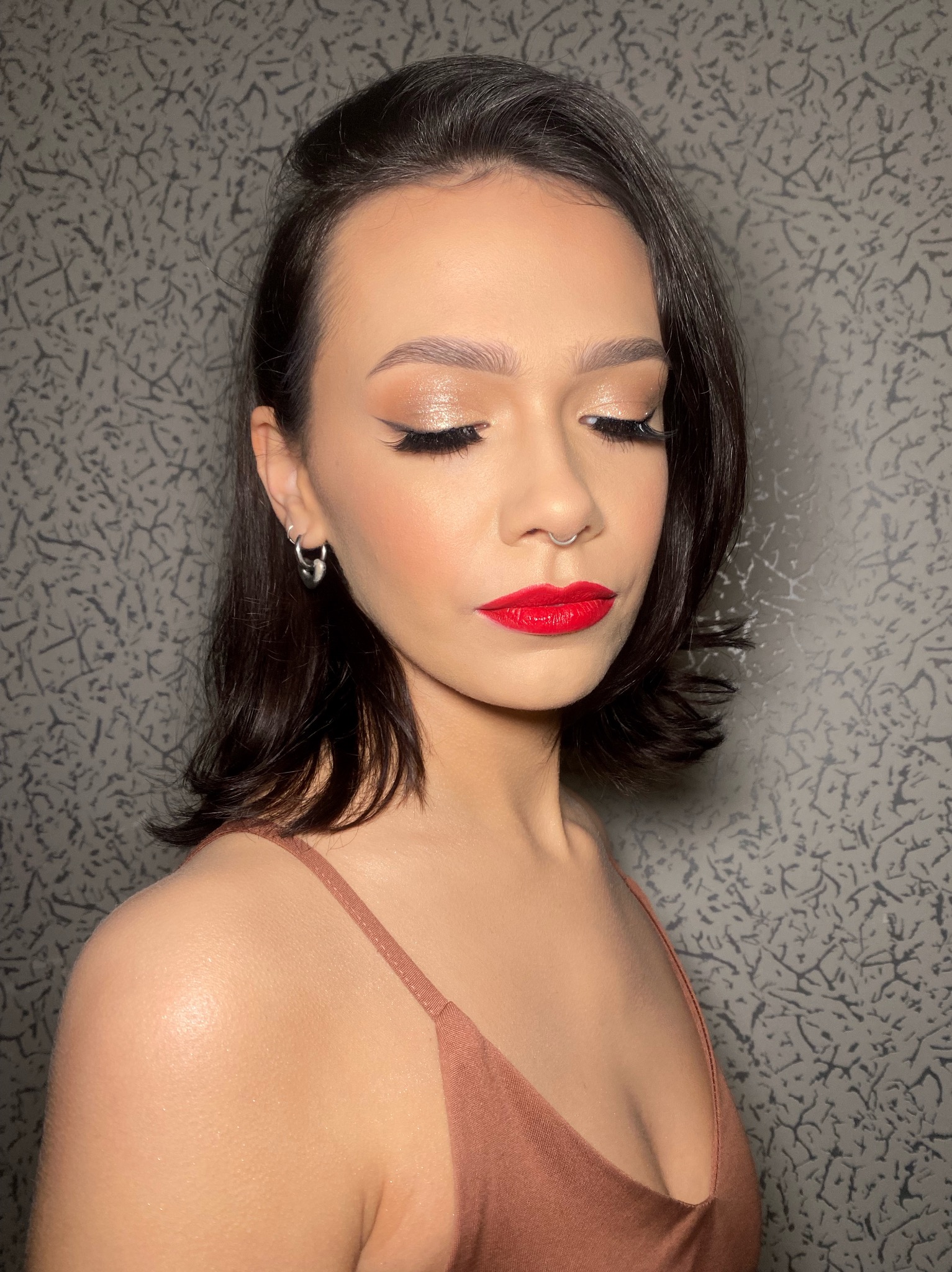 Portret kobiety z zamkniętymi oczami, prezentującej makijaż z błyszczącymi cieniami i intensywnie czerwoną szminką, srebrny kolczyk w uchu i septum.