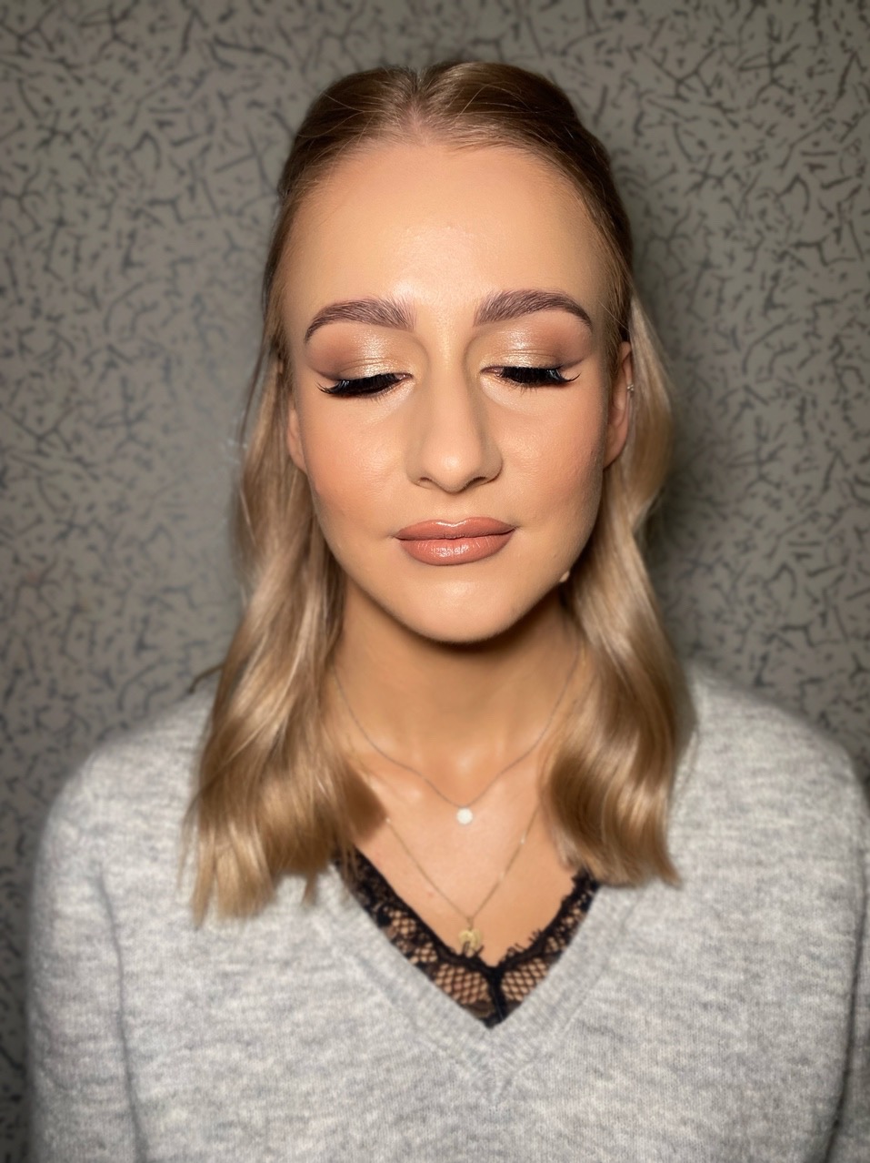 Portret kobiety z zamkniętymi oczami, prezentującej delikatny, złoty odcień na powiekach i precyzyjnie wykonane kreski. Usta w odcieniu nude, subtelna biżuteria.