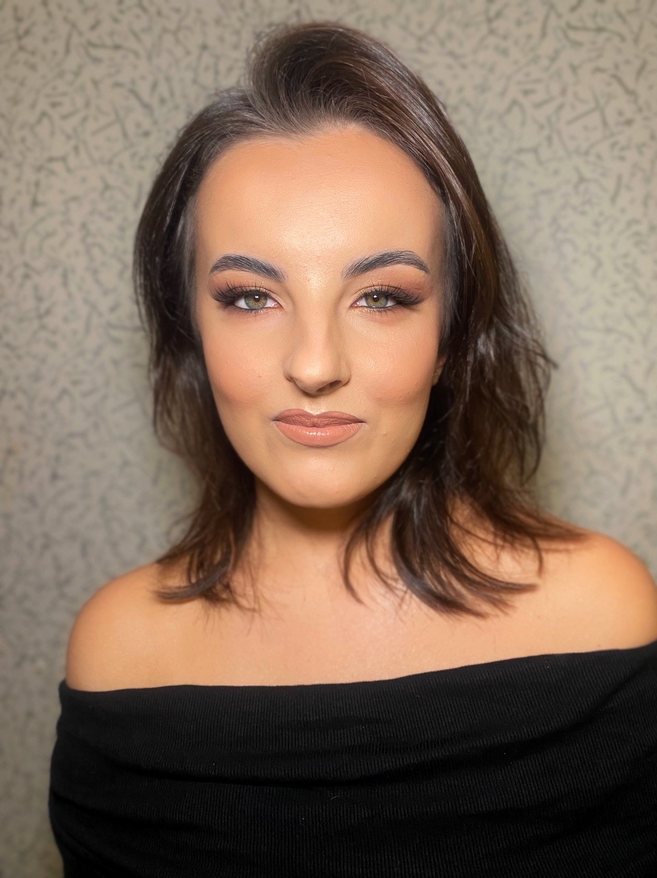 Portret kobiety z brązowymi włosami i profesjonalnym makijażem, z uwzględnieniem cieniowania powiek i podkreślonych ust w odcieniu nude, na tle wzorzystej ściany.