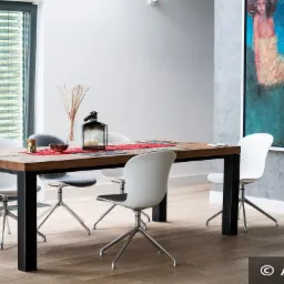 Nowoczesne żaluzje wewnętrzne poziome w szarej ramie okiennej, widoczne za drewnianym stołem z czarnymi nogami i krzesłami w jasnym wnętrzu z obrazem na ścianie.