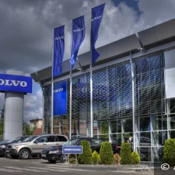 Salon samochodowy Volvo z fasadą osłoniętą zewnętrznymi żaluzjami aluminiowymi, widoczne zaparkowane samochody i firmowe flagi.