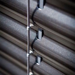 ALUTOR SYSTEMY ROLETOWE S.C. - Szczegółowe ujęcie metalowych listew zewnętrznej osłony okiennej z systemem sznurkowym, widoczne mocowania i faktura materiału.