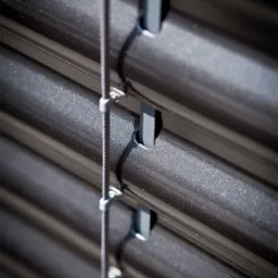 Szczegółowe ujęcie metalowych listew zewnętrznej osłony okiennej z systemem sznurkowym, widoczne mocowania i faktura materiału.