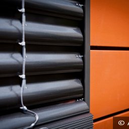 ALUTOR SYSTEMY ROLETOWE S.C. - Detale zamkniętej żaluzji fasadowej w kolorze antracytowym, zamontowanej na elewacji budynku z pomarańczowymi panelami, widoczne prowadnice linkowe.