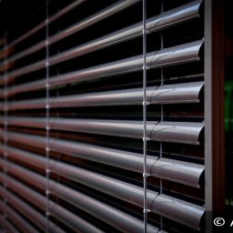 ALUTOR SYSTEMY ROLETOWE S.C. - Zewnętrzne, metaliczne osłony okienne z widoczną strukturą lamel i systemem linek, zamontowane na elewacji budynku z pomarańczowymi elementami.