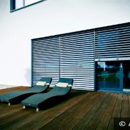 Nowoczesny dom z białymi elewacjami i dużymi oknami osłoniętymi poziomymi lamelami, dwa leżaki na drewnianym tarasie, widok na błękitne niebo z chmurami.