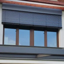 Ciemnoszare poziome lamele okienne zamontowane na elewacji domu z drewnianą ramą okienną i widocznym fragmentem konstrukcji zadaszenia.