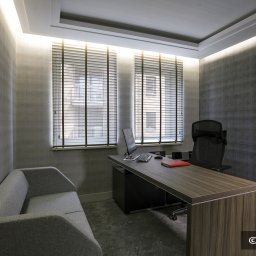 ALUTOR SYSTEMY ROLETOWE S.C. - Biuro z dwoma oknami wyposażonymi w drewniane żaluzje poziome, szarym dywanem, sofą i biurkiem z komputerem.