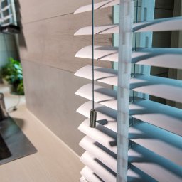 ALUTOR SYSTEMY ROLETOWE S.C. - Szczegółowe ujęcie białych żaluzji z metalowym obciążnikiem sznurka na tle drewnianej ściany i blatu.