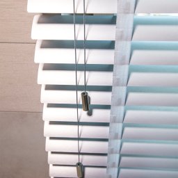 ALUTOR SYSTEMY ROLETOWE S.C. - Szczegółowe ujęcie białych żaluzji drewnianych z metalowymi obciążnikami na sznurkach, widoczne tekstylne taśmy mocujące lamele, neutralne tło ściany.