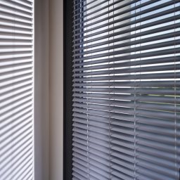ALUTOR SYSTEMY ROLETOWE S.C. - Szare poziome lamele w oknie, rzucające prążkowany cień na ścianę, widok z bliska na regulowane zaciemnienie.