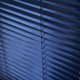 ALUTOR SYSTEMY ROLETOWE S.C. - Ciemne, metalowe lamele żaluzji aluminiowych, ustawione pod kątem, z widocznymi linkami sterującymi i subtelnym światłem prześwitującym między lamelami.