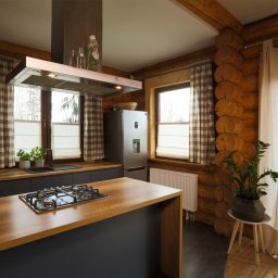 ALUTOR SYSTEMY ROLETOWE S.C. - Wnętrze kuchni w drewnianym domu z widocznymi oknami przesłoniętymi roletami materiałowymi w kolorze białym, drewnianymi blatami i ciemnymi meblami.