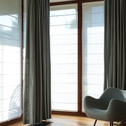 ALUTOR SYSTEMY ROLETOWE S.C. - Nowoczesne wnętrze z dużymi oknami osłoniętymi jasnymi żaluzjami panelowymi i zasłonami w kolorze gołębim, uzupełnione fotelem w stylu retro na drewnianej podłodze w promieniach słońca.