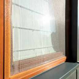 Drewniana rama okienna z siatką przeciwsłoneczną, widoczne poziome listwy i odbicie słońca.