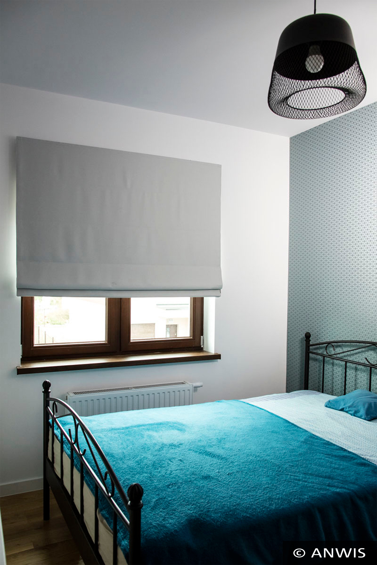 Szara, płaska osłona okienna w stylu rzymskim, zamontowana w brązowym oknie w sypialni z turkusowym kocem na łóżku i ozdobną lampą wiszącą.