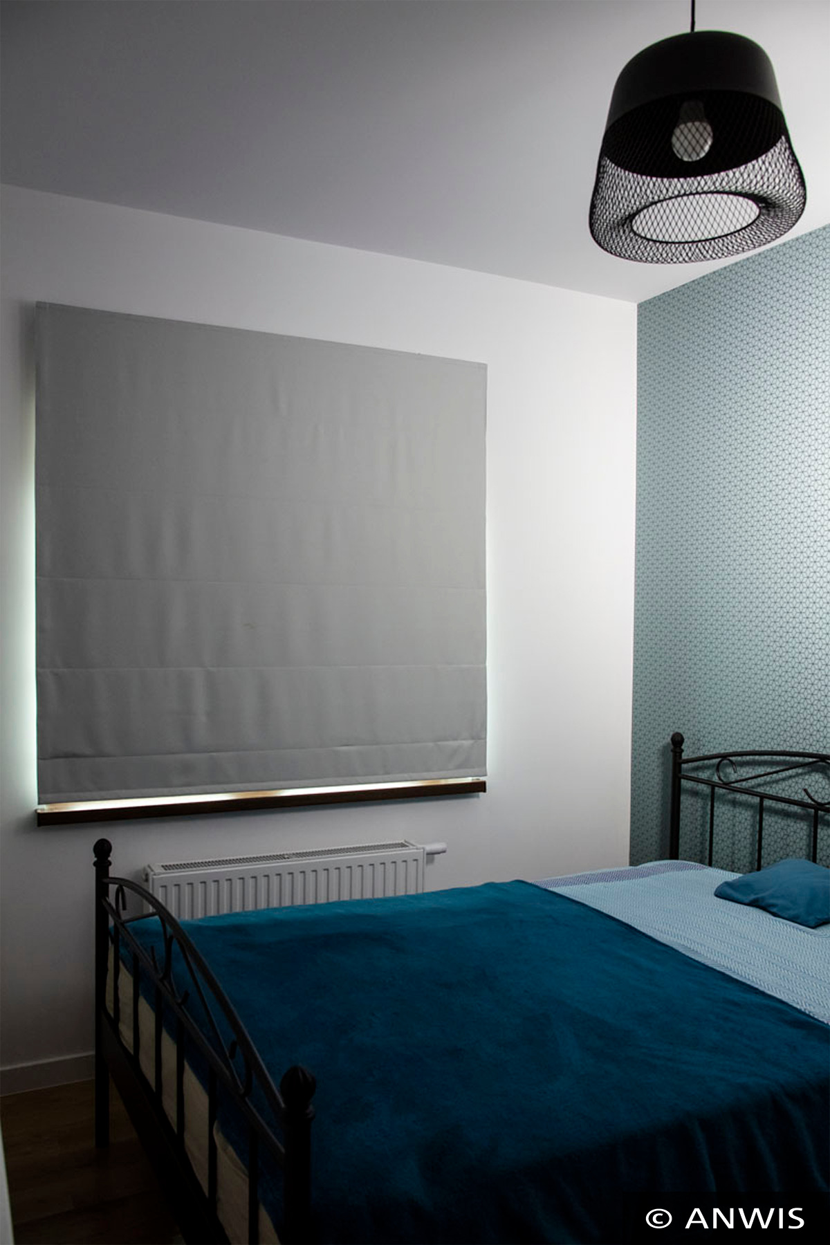 Szara, zwinięta tkanina okienna w stylu rzymskim, zamontowana w sypialni z niebieską tapetą i czarną metalową lampą wiszącą.