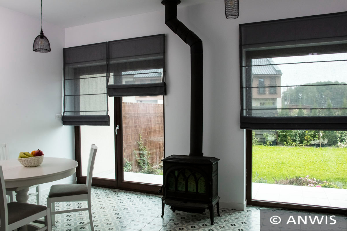 Jasne wnętrze z dwoma oknami przesłoniętymi szarymi roletami rzymskimi, widok na ogród, żeliwny piec kominkowy z czarną rurą, okrągły stół z owocami.
