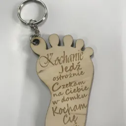 Drewniany breloczek w kształcie stopy z wygrawerowanym tekstem 'Kochanie Jedź ostrożnie Czekam na Ciebie w domku Kocham Cię Twoja walentynka', zawieszony na metalowym kółku.