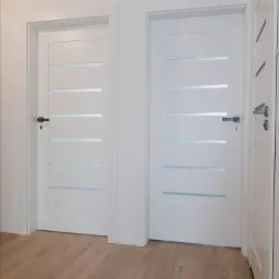 Dwa białe skrzydła z poziomymi, matowymi przeszkleniami, zamontowane w białych ościeżnicach, na podłodze z paneli w odcieniach brązu.