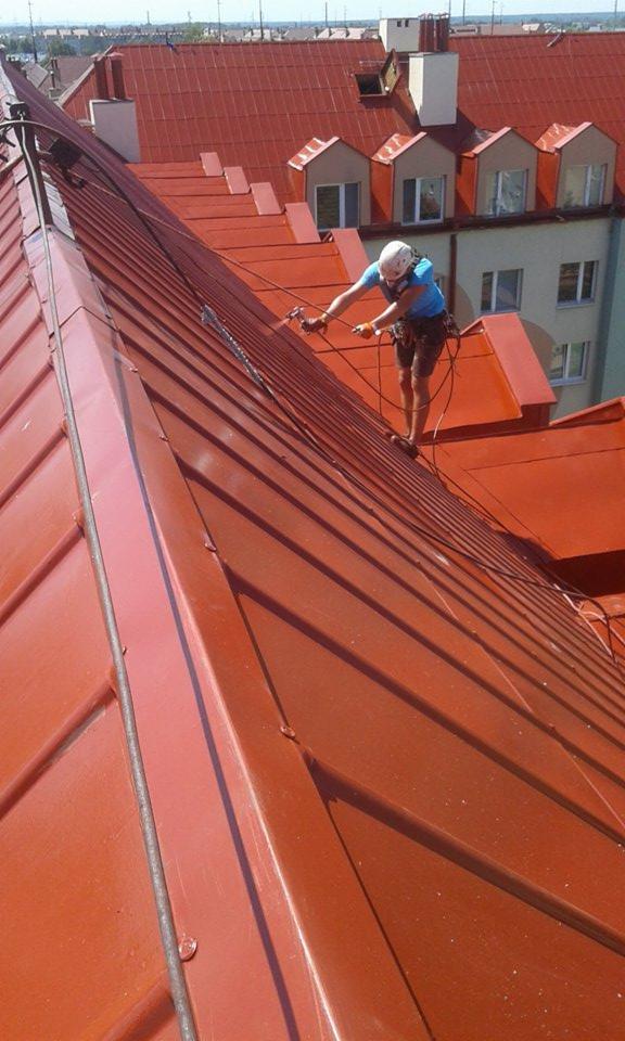 Pracownik w uprzęży, z bliska, maluje czerwoną blachodachówkę na stromym dachu, używając pistoletu natryskowego; w tle kamienica z oknami.