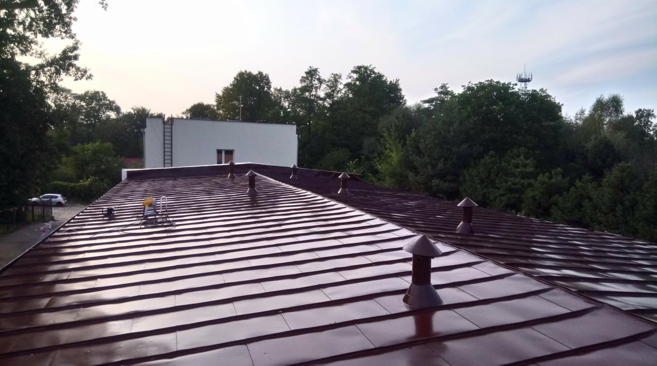 Błyszczący, brązowy dach z blachy trapezowej z kilkoma kominkami wentylacyjnymi; na dachu widoczne narzędzia dekarskie, w tle budynek i drzewa.