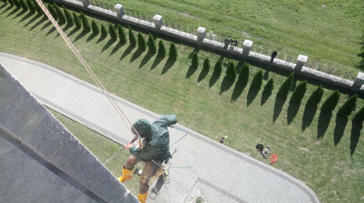 Pracownik w uprzęży, z żółtymi butami, zjeżdża po linie z wysokiego budynku, widok z góry na chodnik z kostki, trawnik z rzędem tuj i fragmentem ogrodzenia.