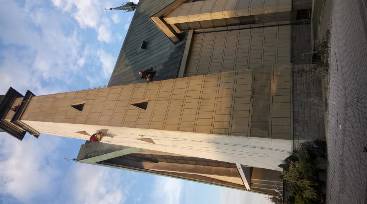 Dwóch alpinistów przemysłowych na linach zabezpieczających, pracujących na wysokości na elewacji kościoła z betonu architektonicznego, widok z dołu na błękitne niebo.