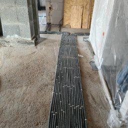 Dabel - Instalacje Elektryczne - Równolegle ułożone rury karbowane w szarym kolorze, prowadzące po surowej betonowej podłodze wewnątrz budynku w trakcie budowy, z widocznymi ścianami z bloczków i płyt OSB w tle.