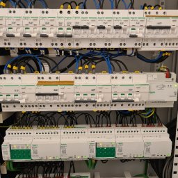 Dabel - Instalacje Elektryczne - Rozdzielnica elektryczna z zainstalowanymi bezpiecznikami i modułami, widoczne przewody w kolorach niebieskim, czarnym i żółto-zielonym, etykiety opisujące obwody, urządzenia marki Schneider