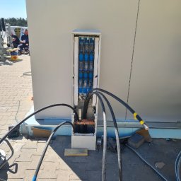 Dabel - Instalacje Elektryczne - Otwarta skrzynka elektryczna na zewnątrz budynku z grubymi kablami wchodzącymi do środka, widoczne bezpieczniki i złącza, na ziemi leżą fragmenty drewna i inne narzędzia.
