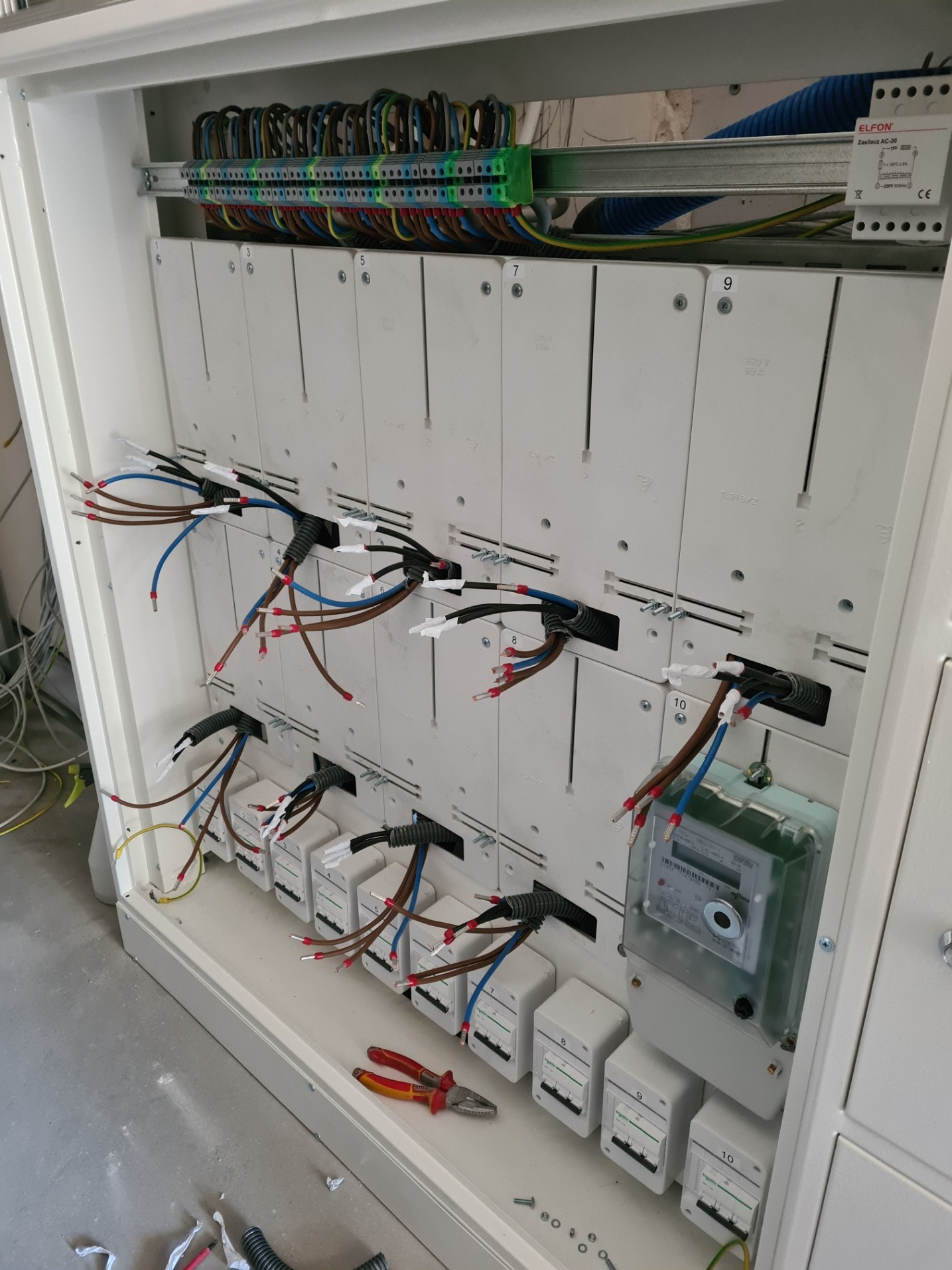 Rozdzielnia elektryczna w trakcie montażu, widoczne podłączone przewody, zabezpieczenia modułowe, licznik energii elektrycznej oraz narzędzia.