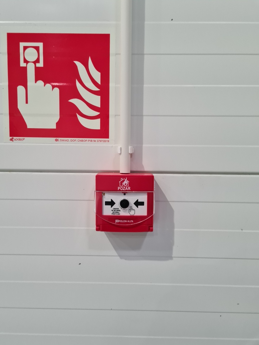 Czerwony, ręczny ostrzegacz pożarowy Polon-Alfa, zamontowany na białej ścianie z poziomymi panelami, obok tabliczka informacyjna z symbolem dłoni naciskającej przycisk i płomieni.