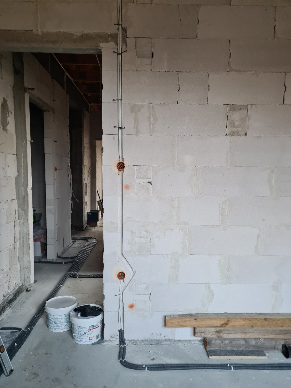 Surowy stan: Wnętrze w trakcie instalacji elektrycznej, widoczne przewody w peszlach prowadzone po ścianie z bloczków, puszki instalacyjne w ścianie, dwa wiadra z farbą i poziomica w tle.