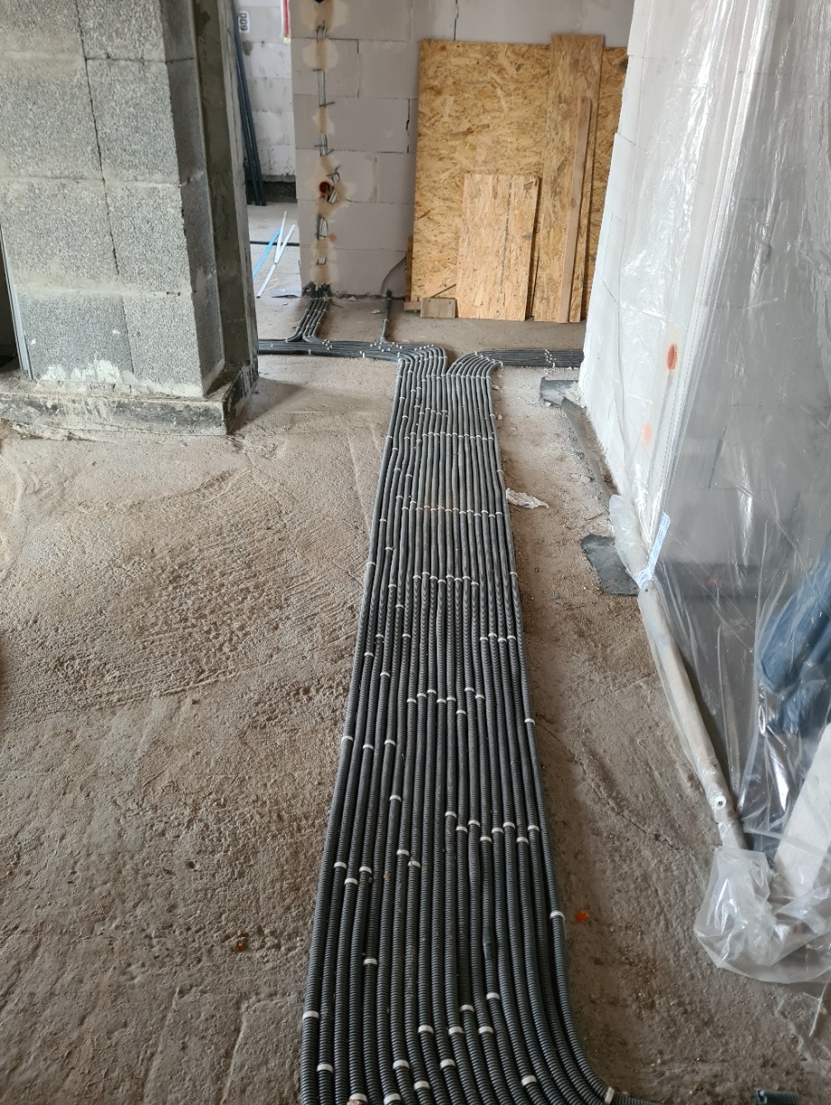 Równolegle ułożone rury karbowane w szarym kolorze, prowadzące po surowej betonowej podłodze wewnątrz budynku w trakcie budowy, z widocznymi ścianami z bloczków i płyt OSB w tle.