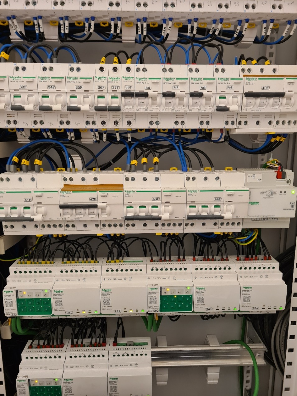 Rozdzielnica elektryczna z zainstalowanymi bezpiecznikami i modułami, widoczne przewody w kolorach niebieskim, czarnym i żółto-zielonym, etykiety opisujące obwody, urządzenia marki Schneider