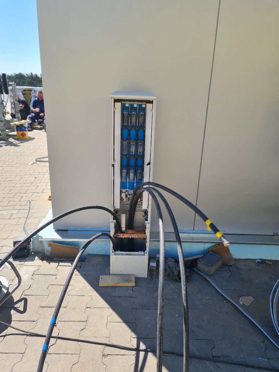 Otwarta skrzynka elektryczna na zewnątrz budynku z grubymi kablami wchodzącymi do środka, widoczne bezpieczniki i złącza, na ziemi leżą fragmenty drewna i inne narzędzia.