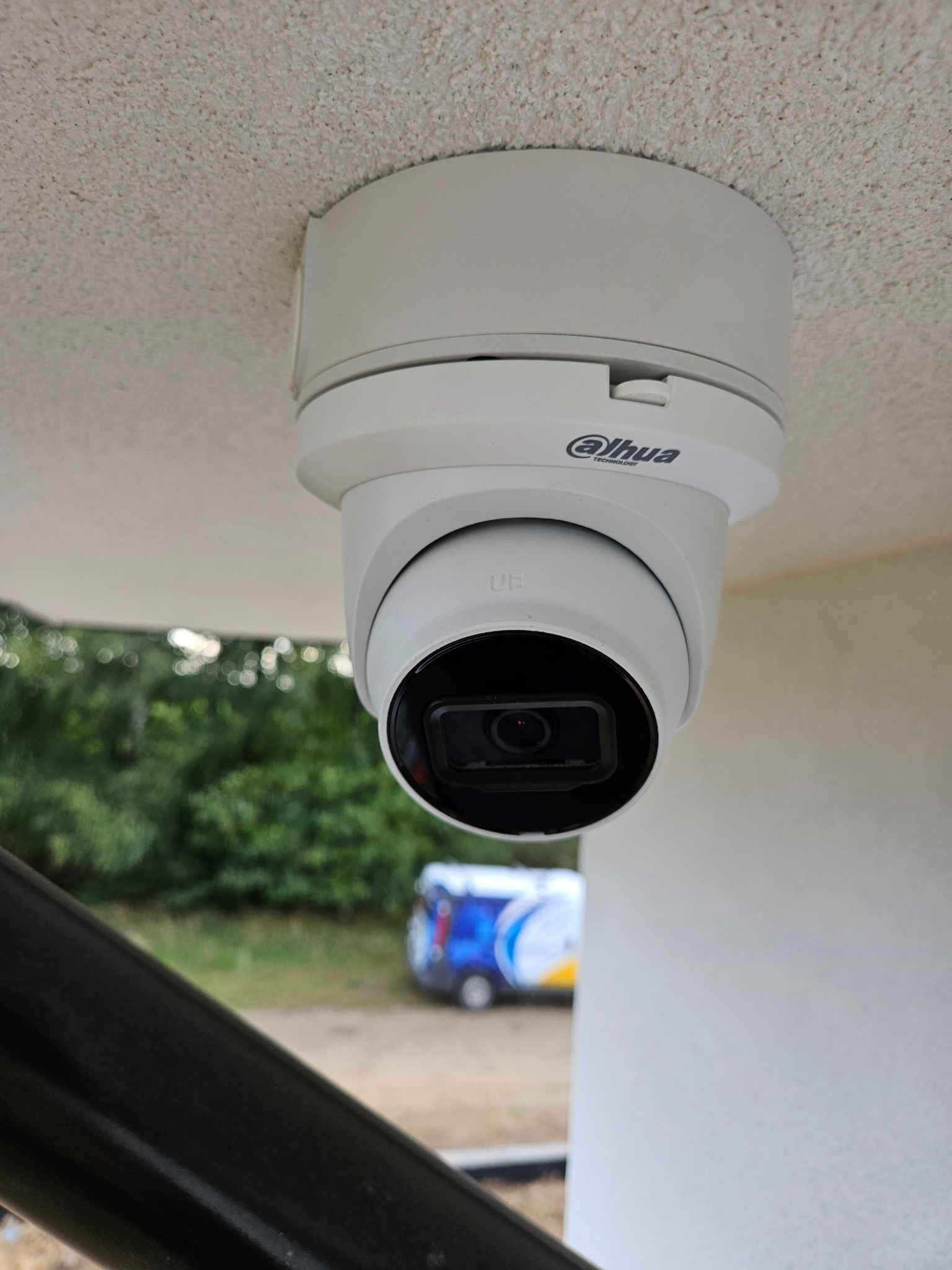 Kamera monitoringu Dahua zamontowana pod sufitem z widocznym logo producenta, z zewnątrz widoczne drzewa i pojazd z reklamą.