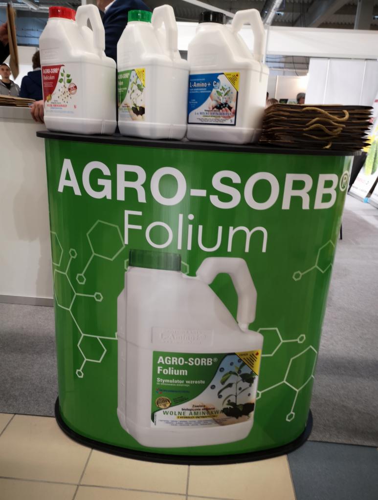 Prezentacja produktów Agro-Sorb Folium na stoisku targowym, widoczne białe kanistry z etykietami i zielony panel z logo firmy.