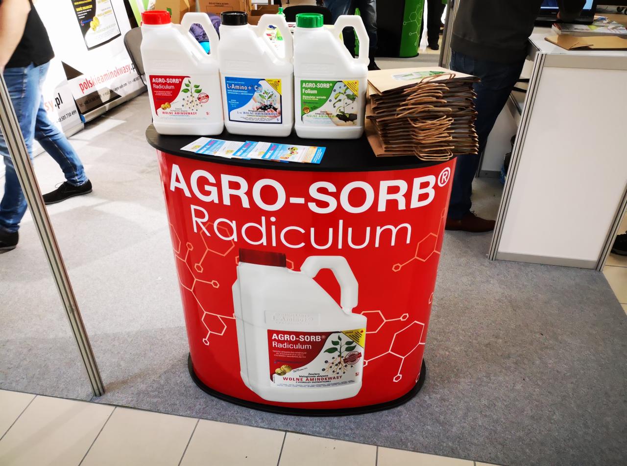 Ekspozycja handlowa w Lublinie prezentująca trzy białe kanistry z etykietami różnych rodzajów preparatów na tle stoiska z logo Agro-Sorb Radiculum, obok leżą ulotki i papierowe torby.