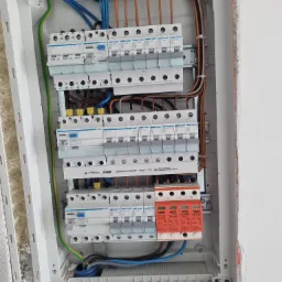 Otwarta skrzynka elektryczna z widocznymi bezpiecznikami, przewodami różnego koloru i zabezpieczeniami przeciwprzepięciowymi VCX, zamontowana w ścianie z surowym wykończeniem.